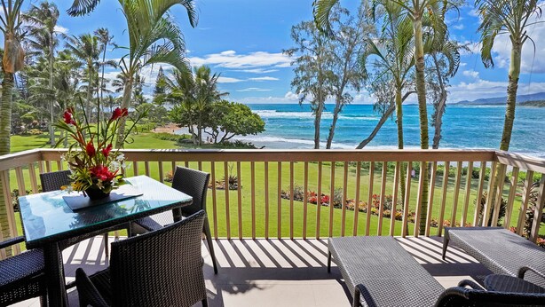 Un balcon avec une table en verre, des chaises en osier et une vue sur l’océan et les palmiers.