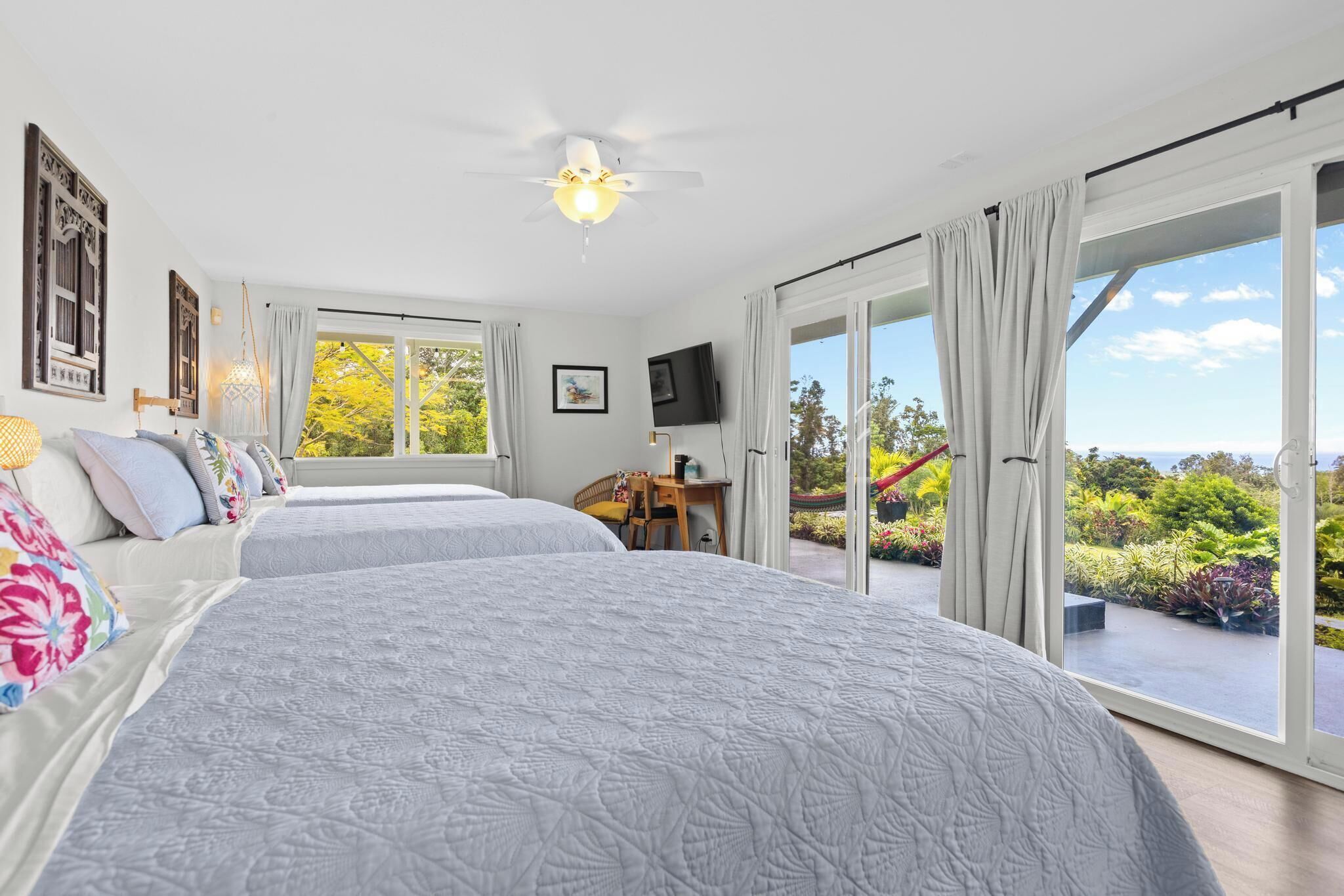 A bedroom with two beds, a ceiling fan, a TV, and a balcony with a view of greenery.