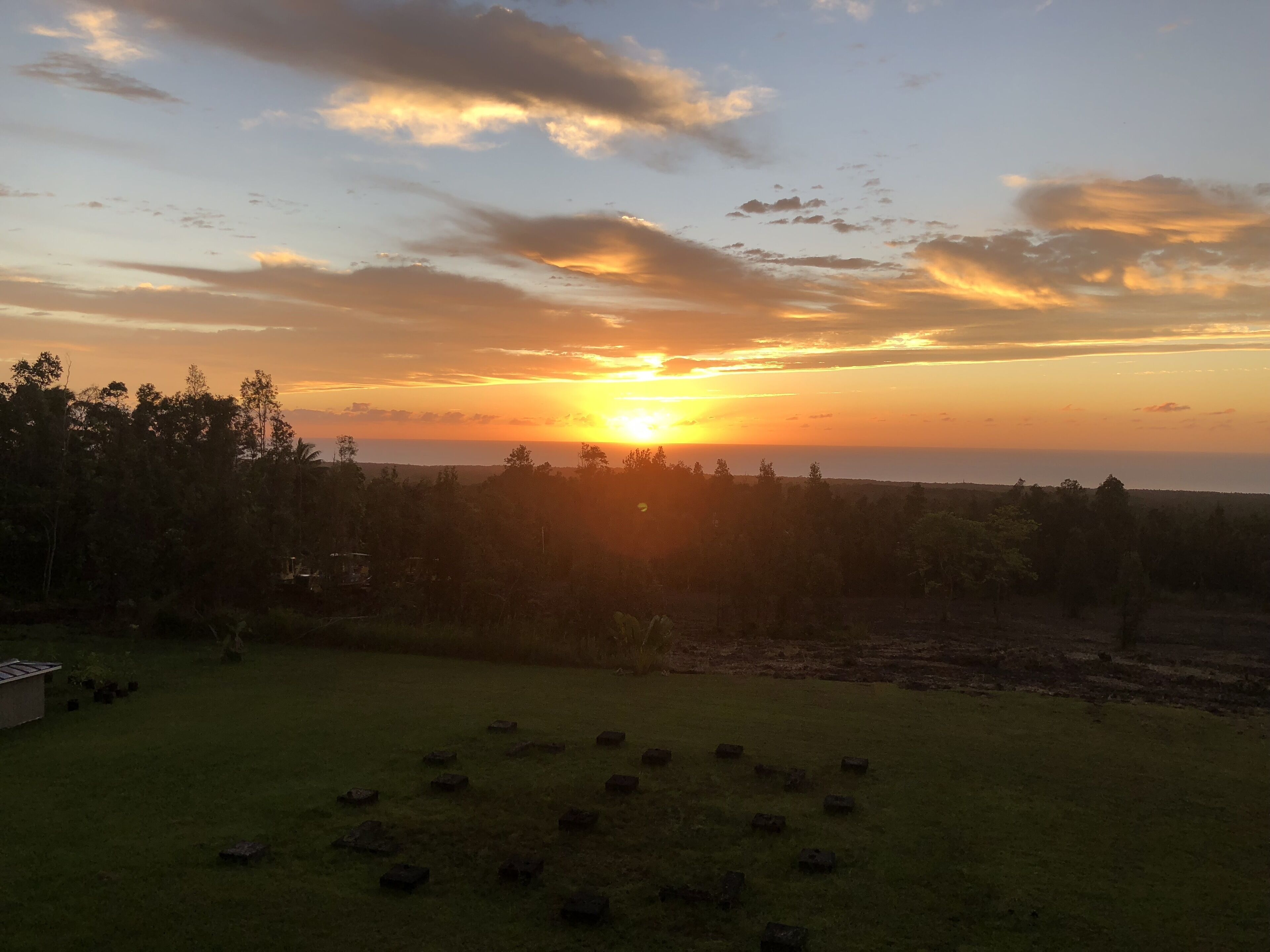 A sunset over a grassy field with scattered trees and a building in the foreground.