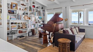 A living room with a grand piano, bookshelves, a sofa, and a view of the ocean. - Mala o Kilauea Nature Retreat; Pool Spa, Ocean views, Orchard, Private and Gated (Pahoa)