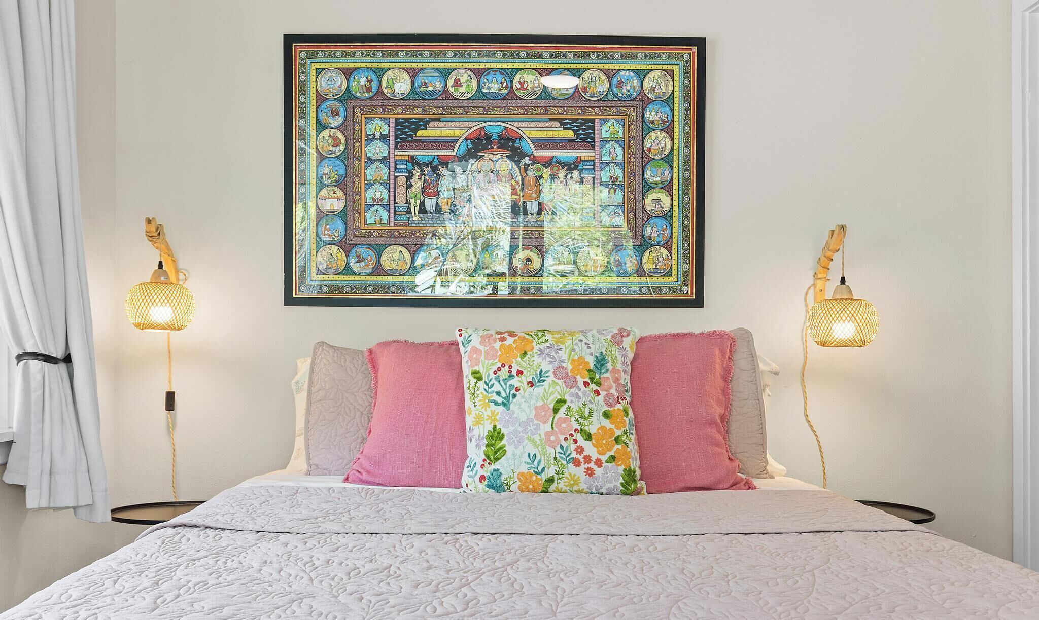 A bed with a white quilt, pink and beige pillows, and a colorful floral throw. A large framed artwork is centered above the bed.