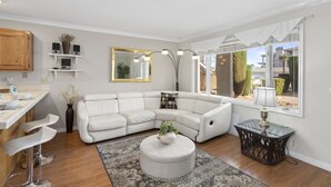 A modern living room with a sectional sofa, a round ottoman, and a glass coffee table. There is a kitchen area with a white countertop and bar stools. A large mirror and pendant lights are visible. - Las Palmas Resort 304, Main Level Condo, New Flooring, Mountain Views, Close to Pools, and more (St. George)