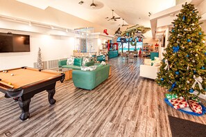 A spacious indoor area with a pool table, green sofas, a Christmas tree, and a view of the ocean through large windows.