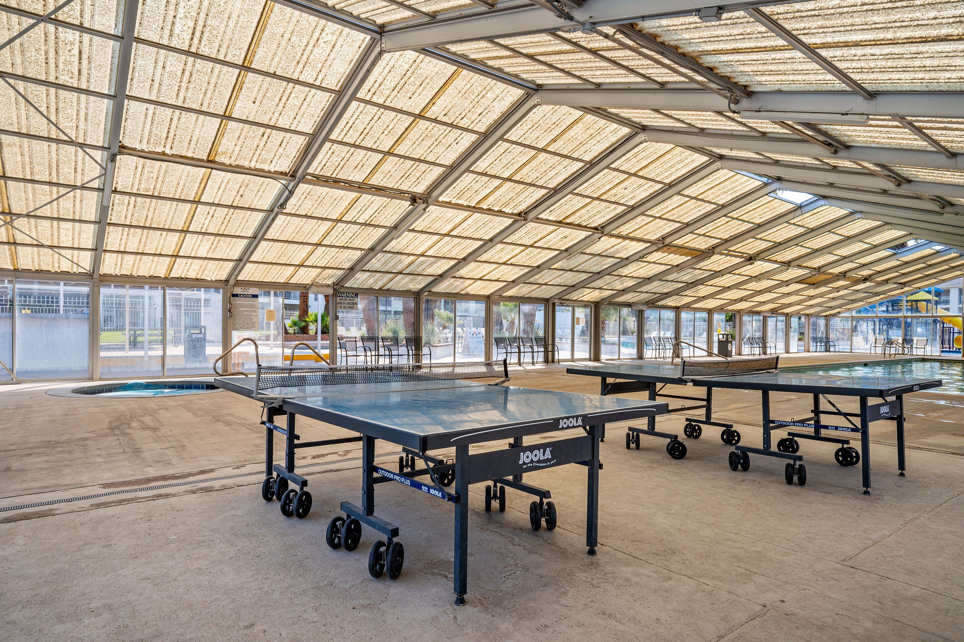 Een sportfaciliteit binnenshuis met meerdere tafeltennistafels, een zwembad en een overdekt gebied met een metalen dak.