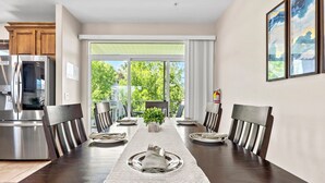 A dining area with a long table set for six, a kitchen with a stainless steel refrigerator, and a view of greenery through large windows. - Las Palmas Resort 1805, Resort Access in St George (St. George)