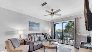 A modern living room with a sofa, armchair, and coffee table, a ceiling fan, and a view to an outdoor patio. - Lakeview #6 | 1BR • Remodeled • Beach Access Across Street ✨ (Miramar Beach)
