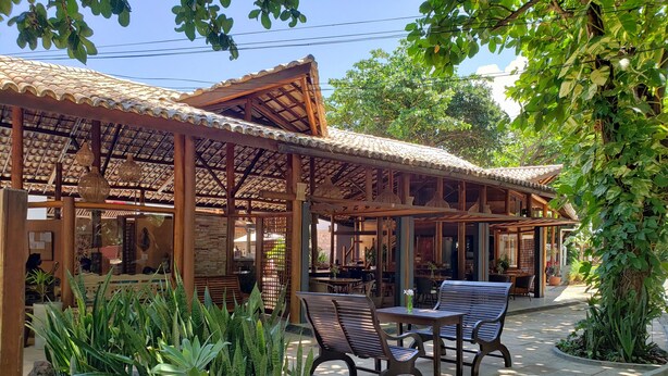 An outdoor seating area with wooden tables and chairs, surrounded by potted plants and a pergola structure. - Pousada Casa de Praia (Mata de São João)