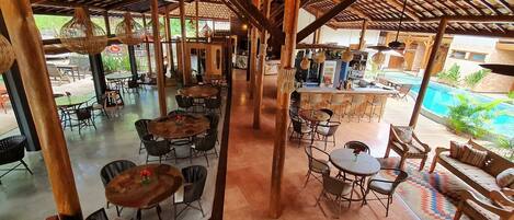 An outdoor restaurant with round tables and chairs, a bar area, and a pool in the background.