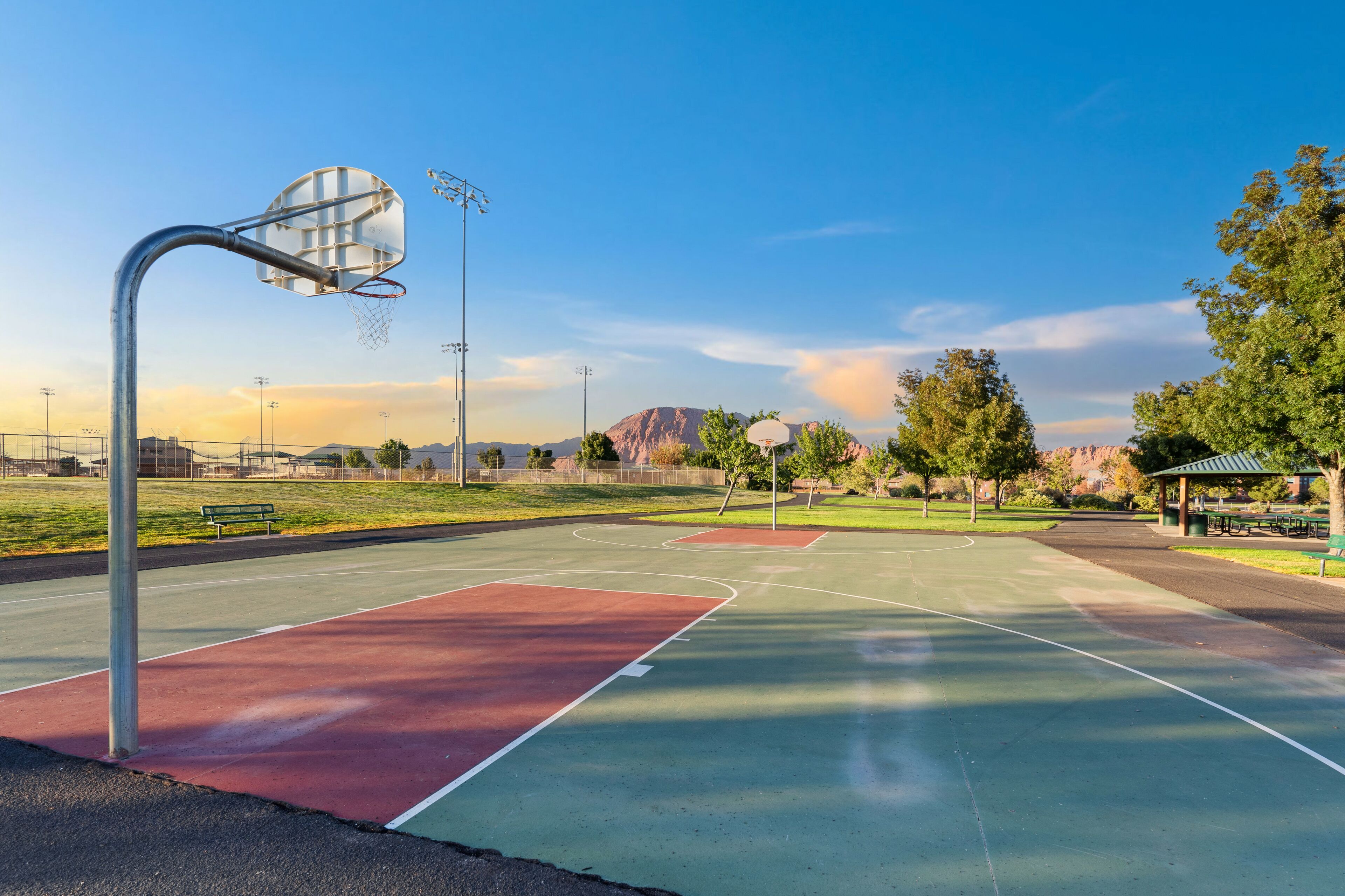 Ein Basketballplatz im Freien mit Metallkorb und -tafel, umgeben von Bäumen und einer Picknickzone.