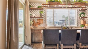 Una cocina con zona de comedor, con armarios de madera, una ventana con persianas y plantas colgantes.