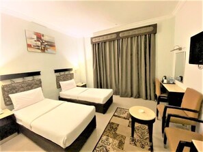 Une chambre d’hôtel avec deux lits, un bureau, une chaise, une petite table et une grande fenêtre avec des rideaux.
