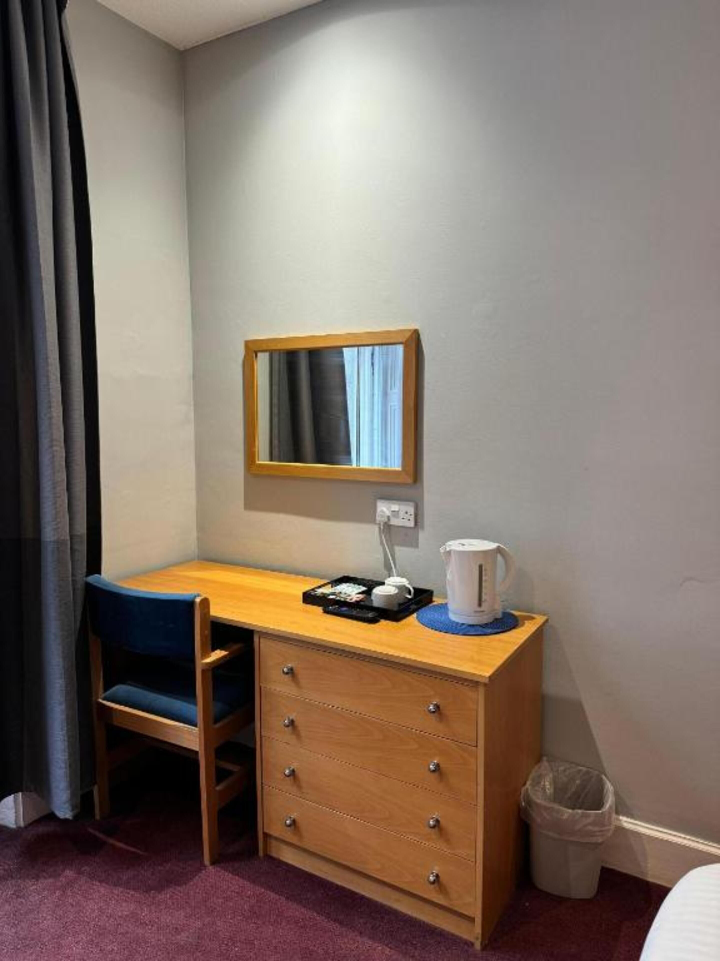 A compact room with a wooden desk, a chair, a mirror, a small trash bin, and a bed.