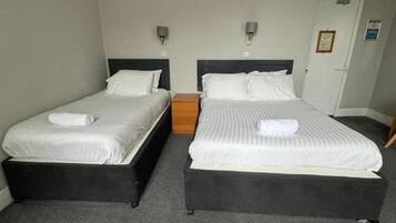 Habitación de hotel con dos camas individuales, cada una con cabecera de madera y colcha blanca. Hay dos mesitas de noche, una en cada cama, y una pequeña mesita auxiliar de madera entre las camas. La habitación tiene alfombra gris y paredes de color claro.