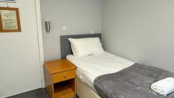 A small, single-bed room with a wooden nightstand, a gray blanket, and a white pillow.