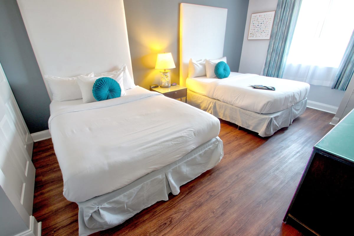 A hotel room with two beds, wooden flooring, a nightstand with a lamp, and a large mirror on the wall.