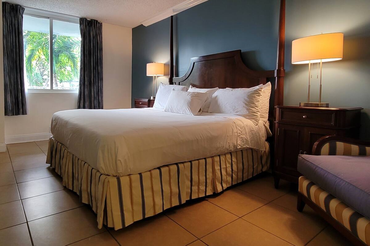 A neatly made bed with a striped skirt, a nightstand with a lamp, a chair with a striped cushion, and a window with curtains.