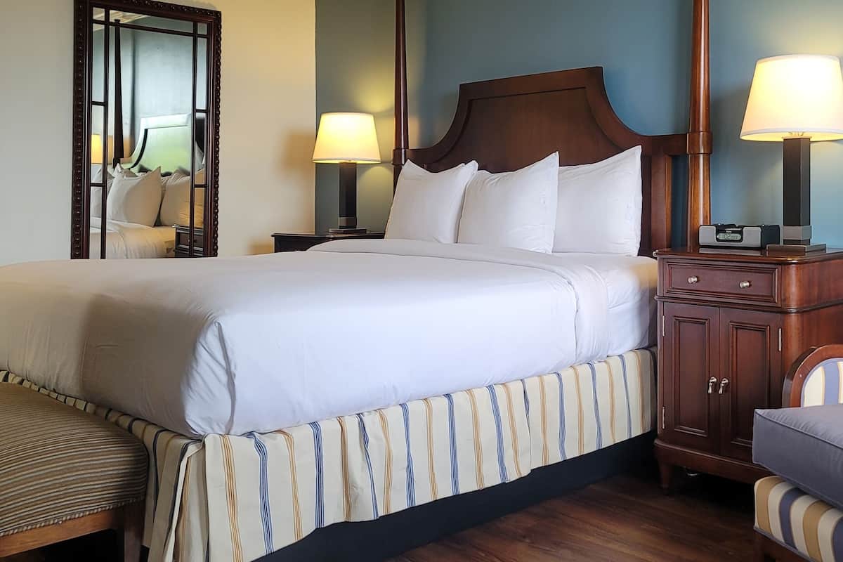 A neatly made bed with white linens, a wooden nightstand with a lamp, a striped chair, and a mirror on the wall.
