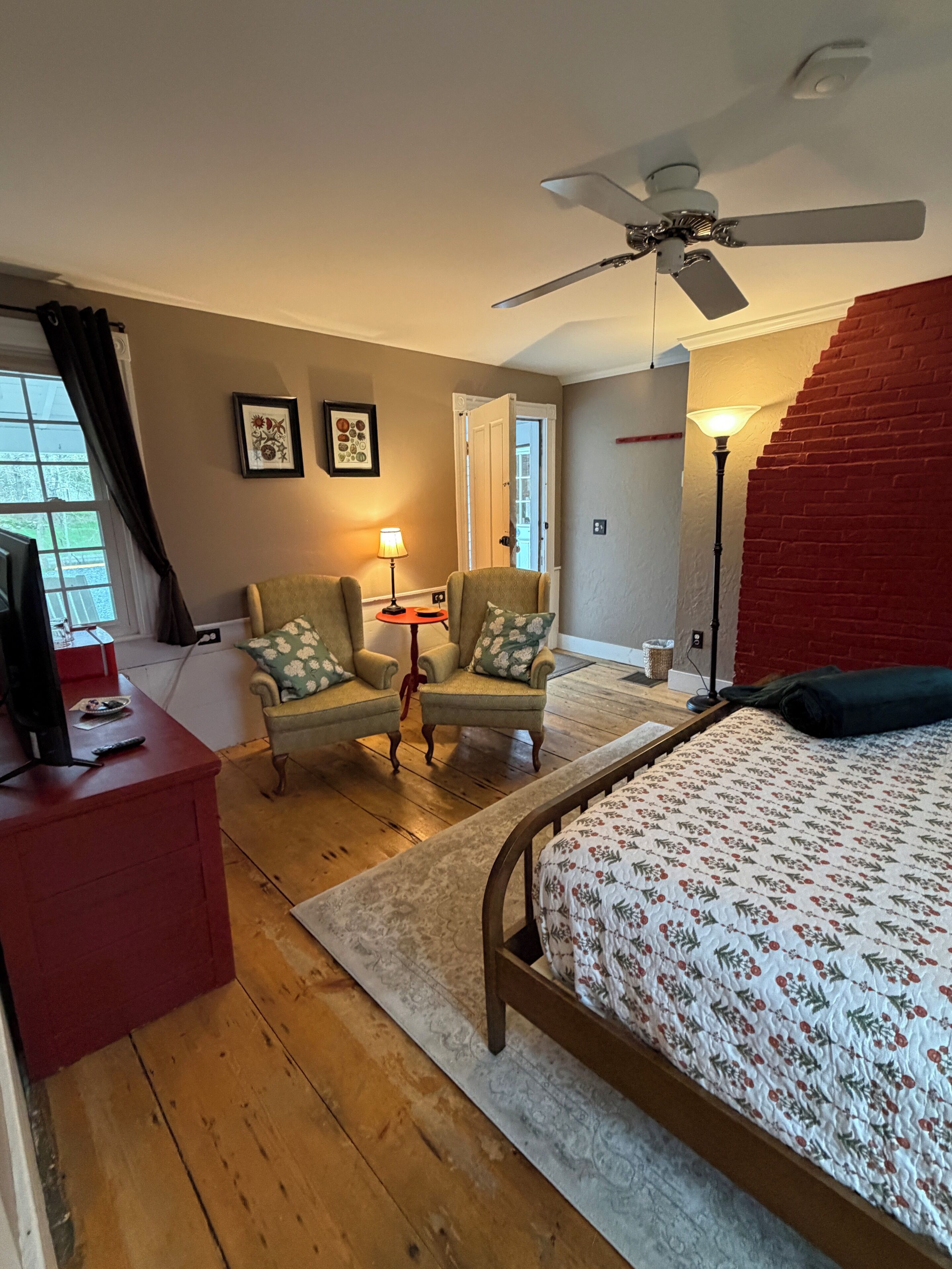 A bedroom with a bed, a chair, a desk, a ceiling fan, and a red brick wall.