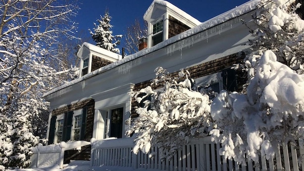 Uma casa coberta de neve com cerca branca de piquete, varanda frontal e árvores carregadas de neve.