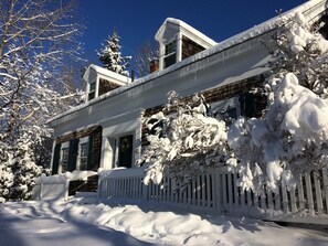 Una casa cubierta de nieve con una valla de piquetes blancos, un porche y árboles cargados de nieve.