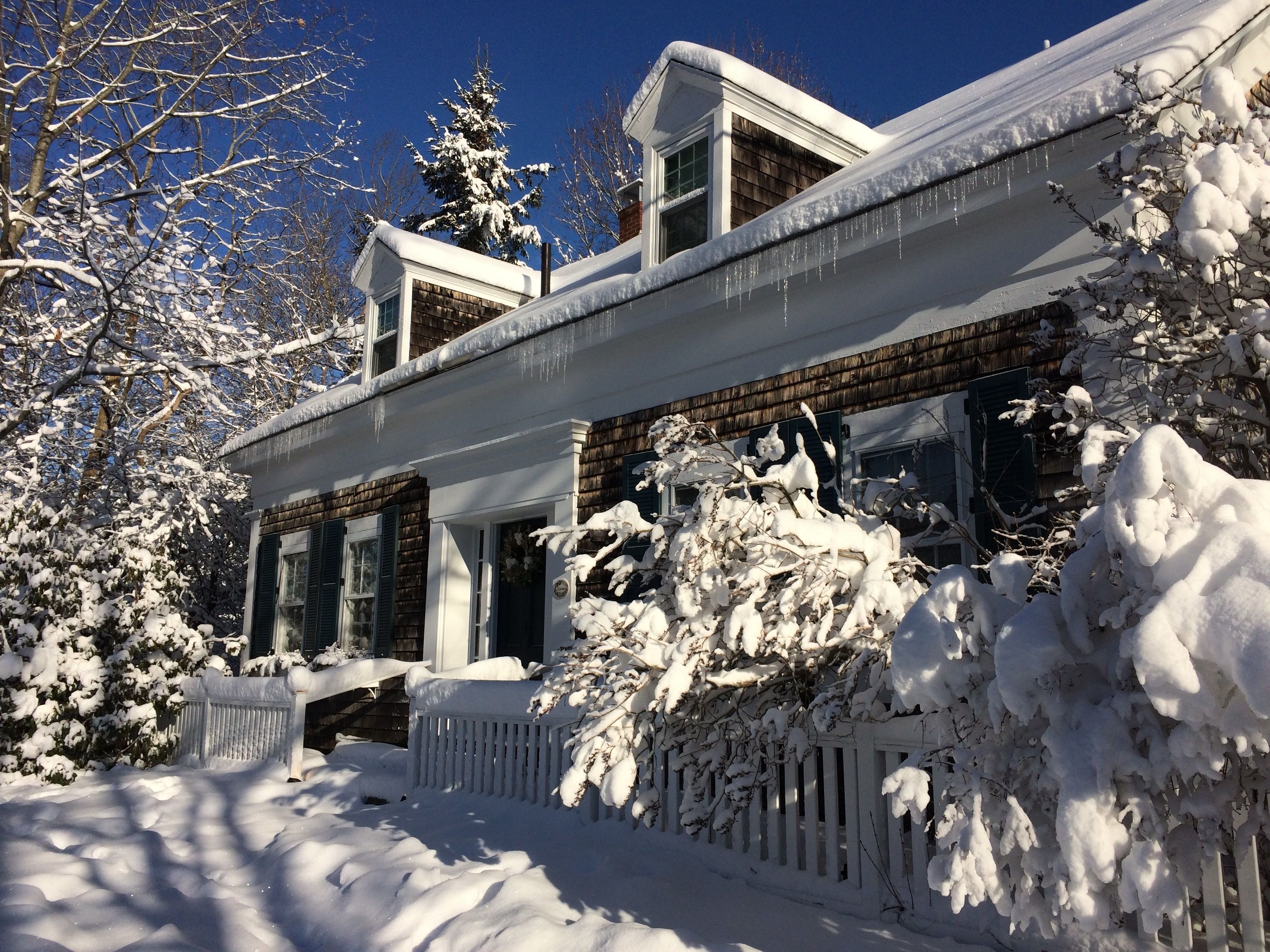 Una casa cubierta de nieve con fachada de madera, una cerca de piquetes blancos y árboles cargados de nieve.