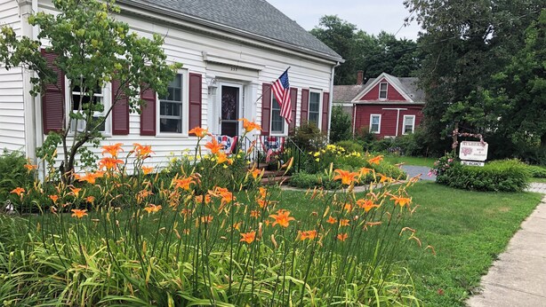 A white house with red shutters and a flag, surrounded by a garden with orange and pink flowers. - Mulberry Bed & Breakfast (Wareham)