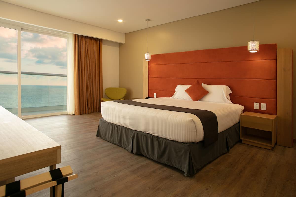 Une chambre d’hôtel moderne avec un grand lit, une table de chevet en bois, une petite table et une vue sur la mer par une grande fenêtre.