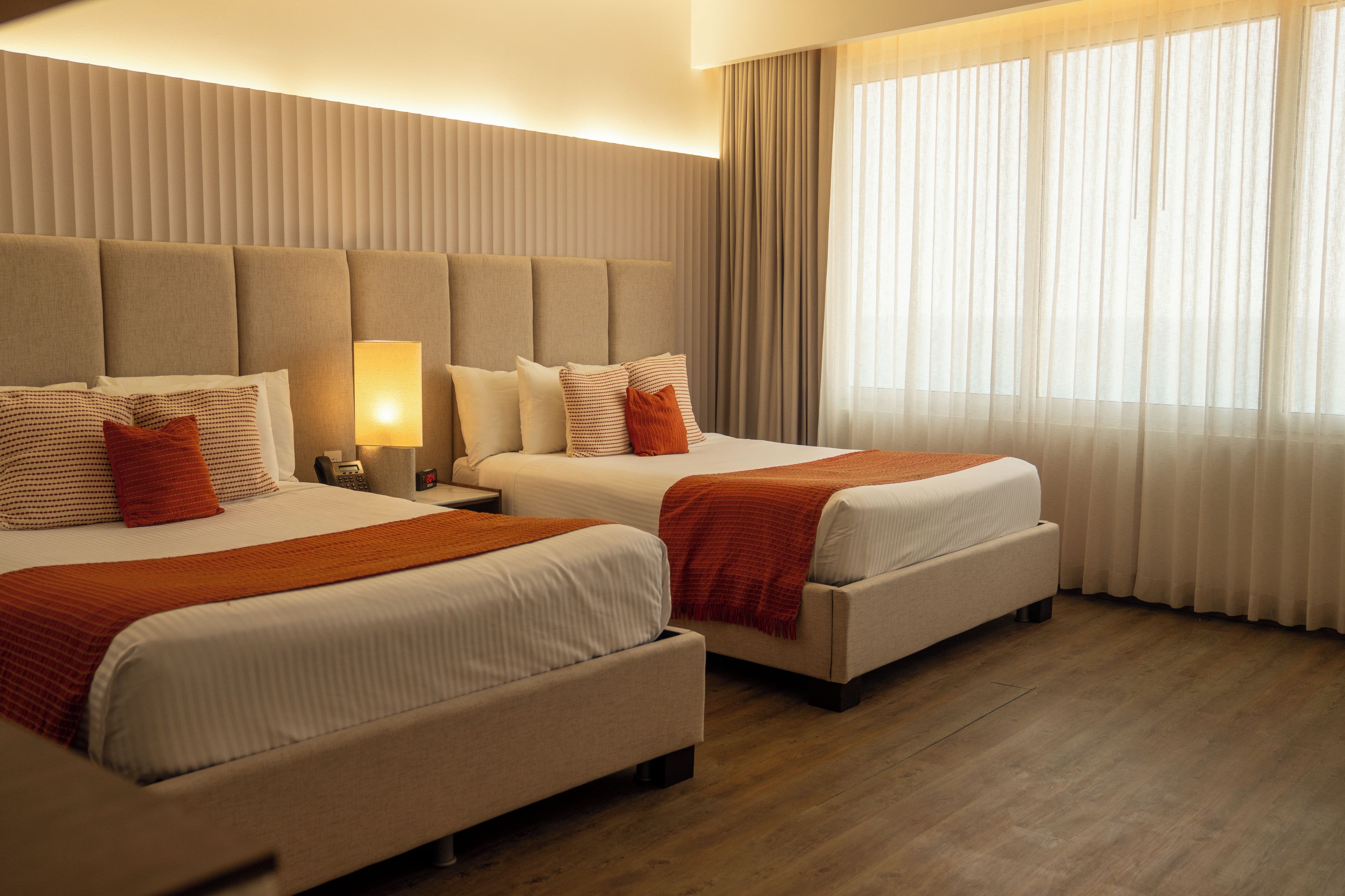 A hotel room with two beds, a nightstand, a lamp, and a window with curtains.