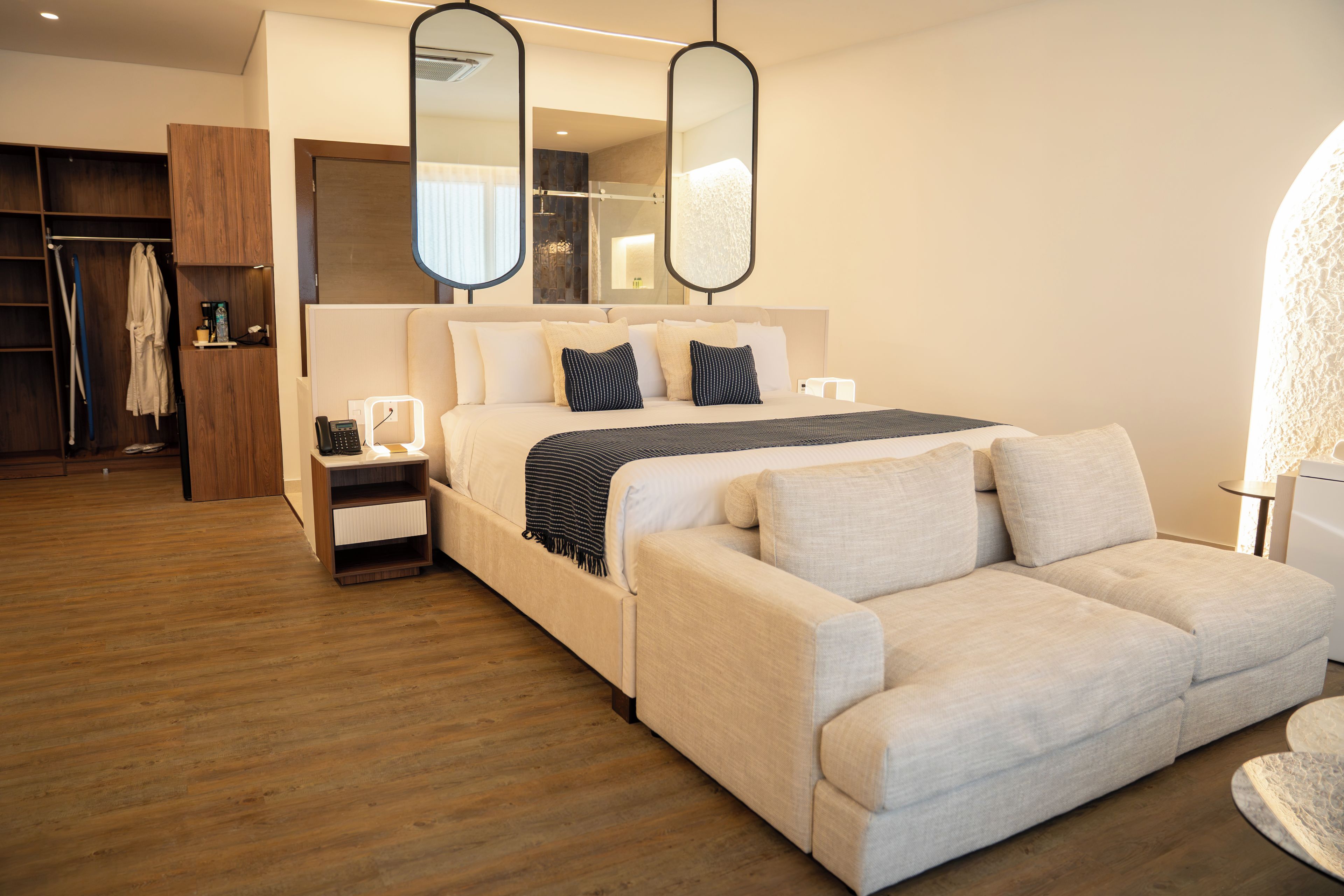 Una habitación de hotel moderna con cama, sofá, mesita de noche, espejo y armario.