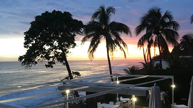 An outdoor dining area with tables and chairs, overlooking the ocean with palm trees and a sunset in the background.