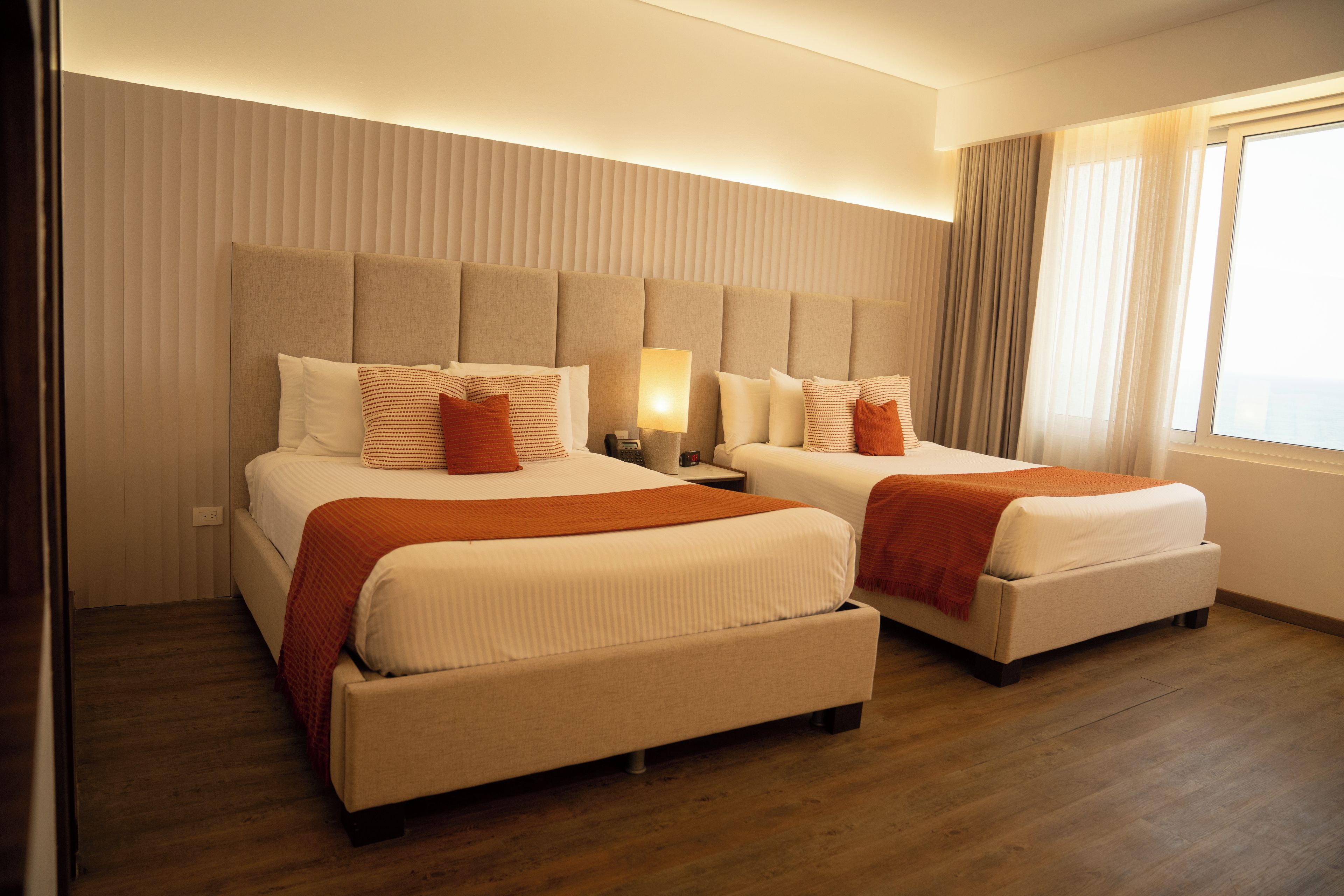 A hotel room with two beds, a nightstand, a lamp, and a window with curtains.