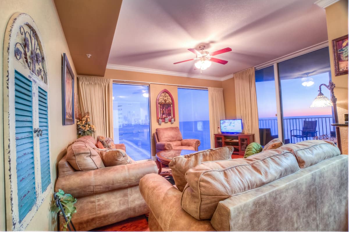 Ein Wohnzimmer mit Ledersofas, einem Deckenventilator, einem Fernseher und Blick auf die Stadt durch große Fenster.