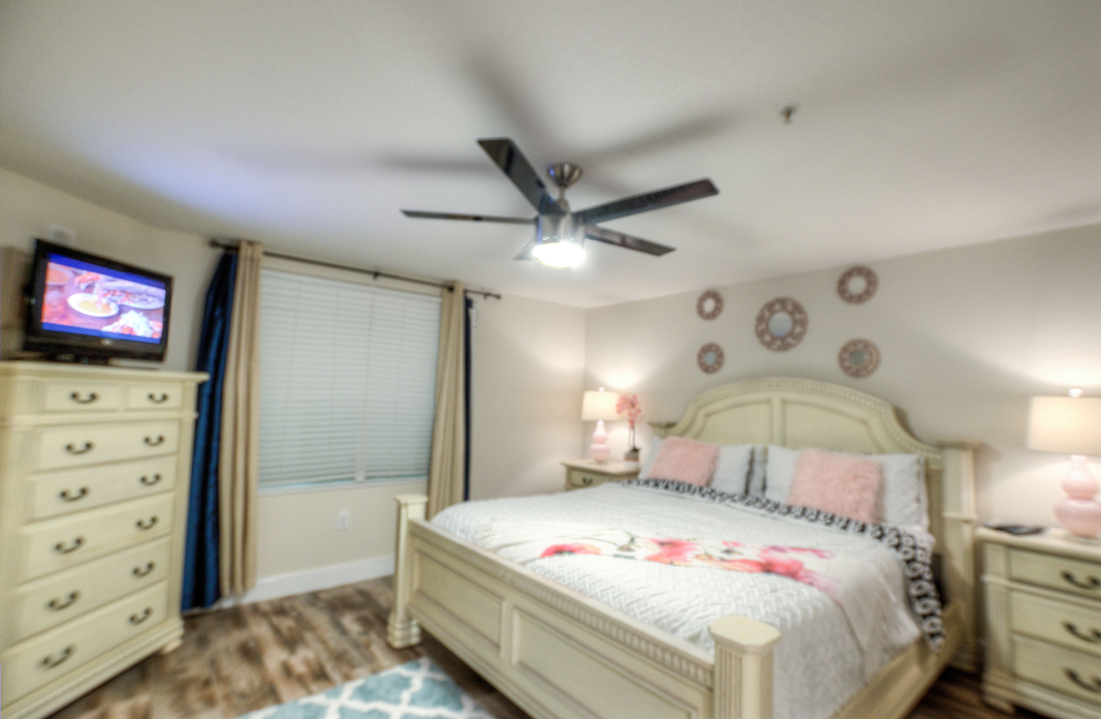 A bedroom with a bed, a ceiling fan, a television, a dresser, and a window with blinds.