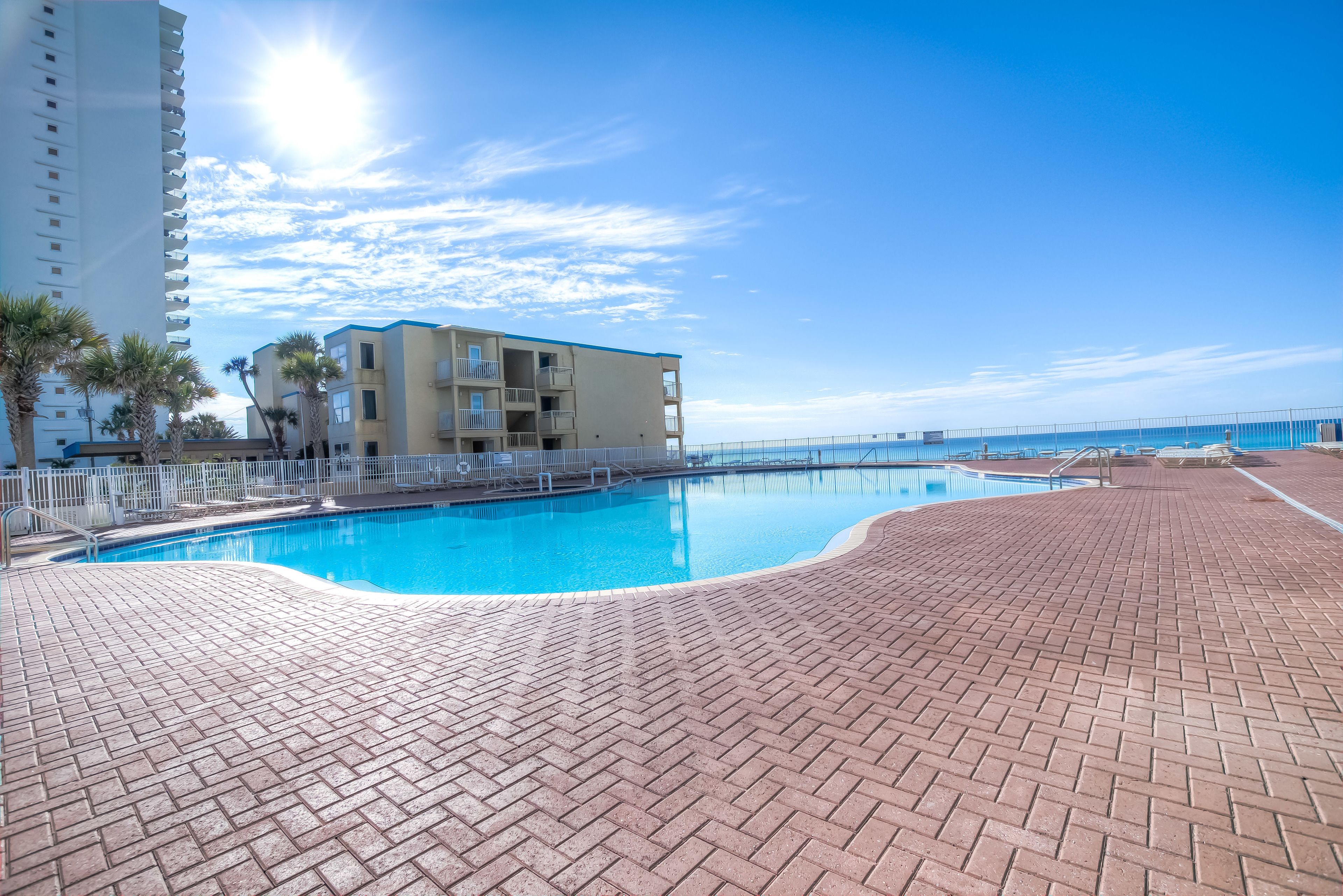 A swimming pool with a brick deck, a building with balconies, and a clear blue sky.