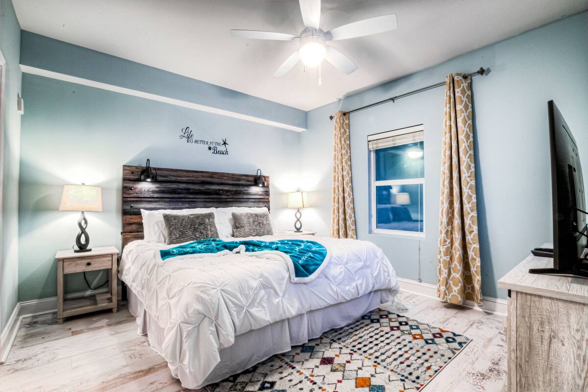 A bedroom with a bed, two bedside tables with lamps, a ceiling fan, a window with curtains, and a television.