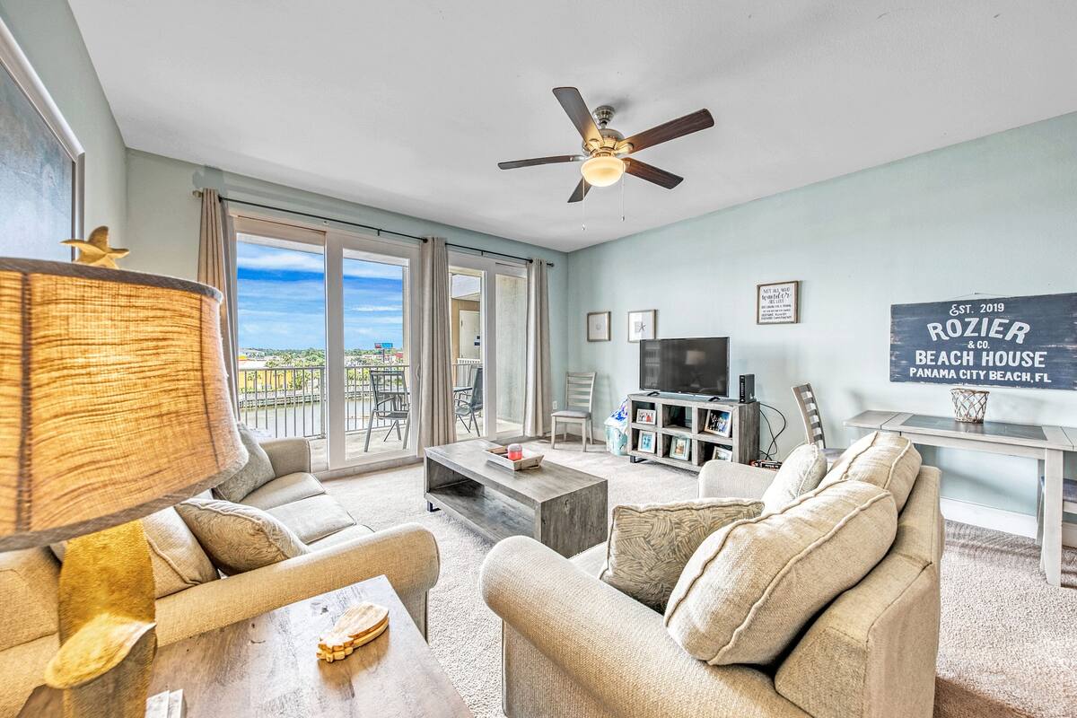 A spacious living room with a large window, a ceiling fan, a flat-screen TV, and a sign indicating the property is a beach house.