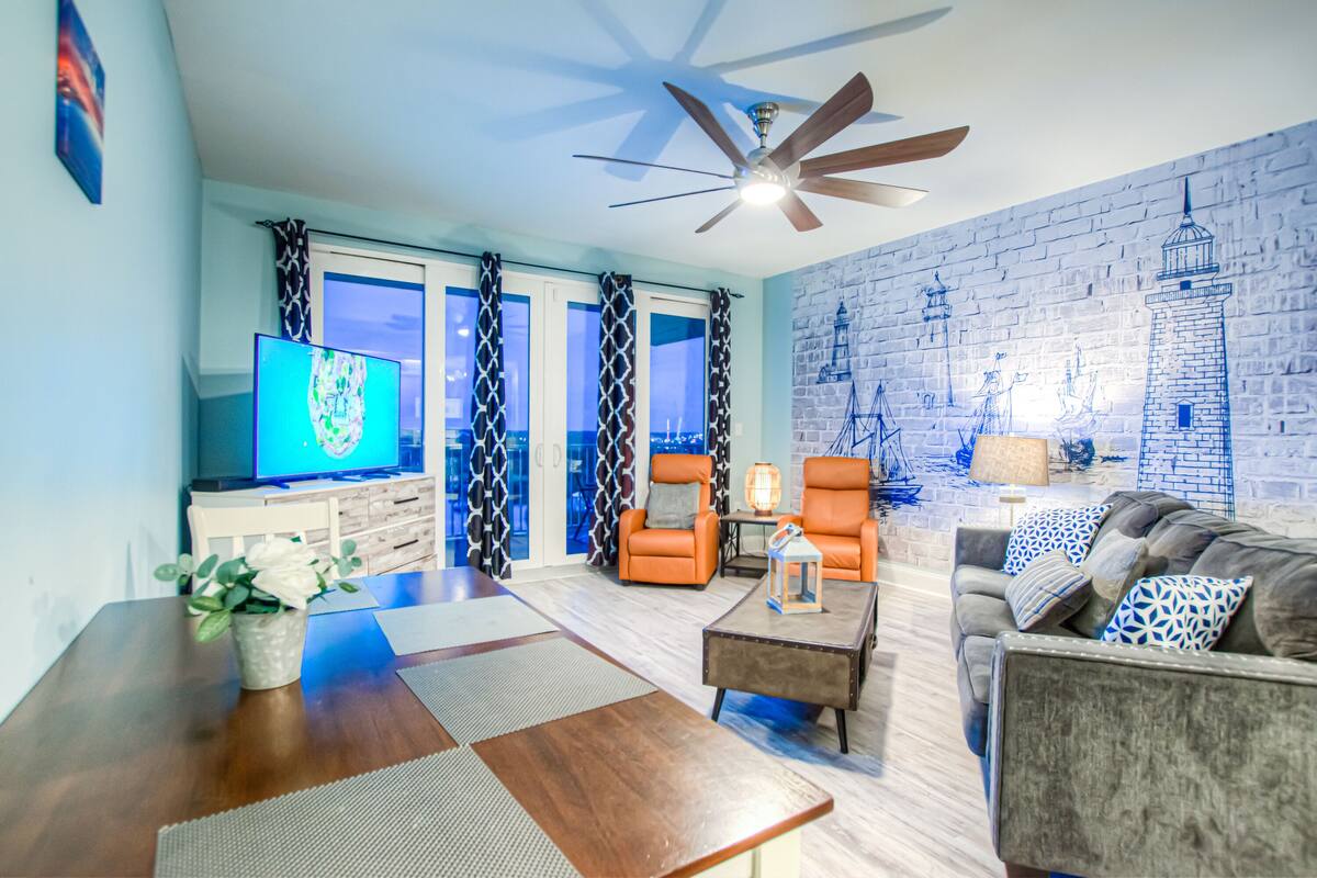 A modern living room with a flat-screen TV, two orange armchairs, a grey sofa, a wooden coffee table, and a brick-patterned wall.