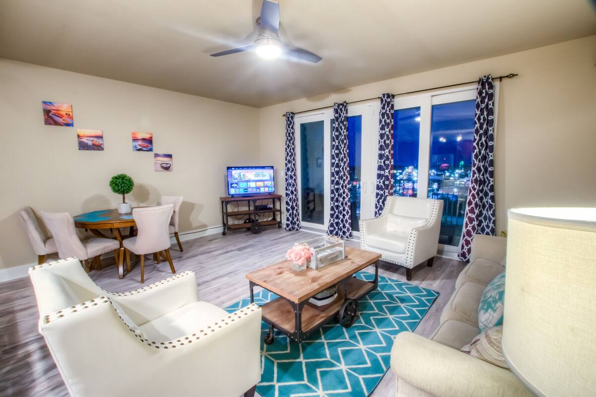 A living room with a TV, white chairs, a coffee table, and a city view through the window.