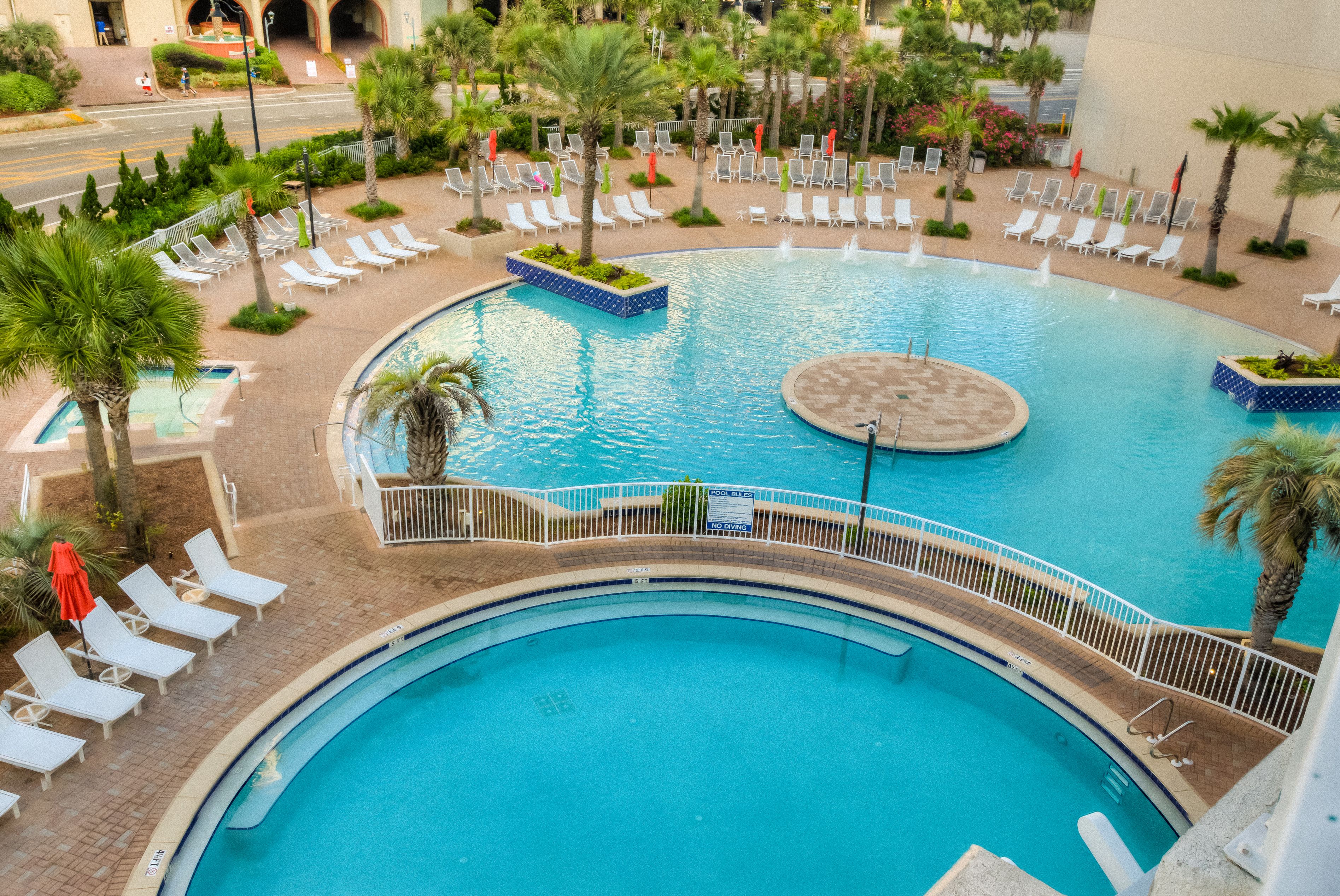A large outdoor swimming pool with multiple lounge areas, palm trees, and a circular water feature.