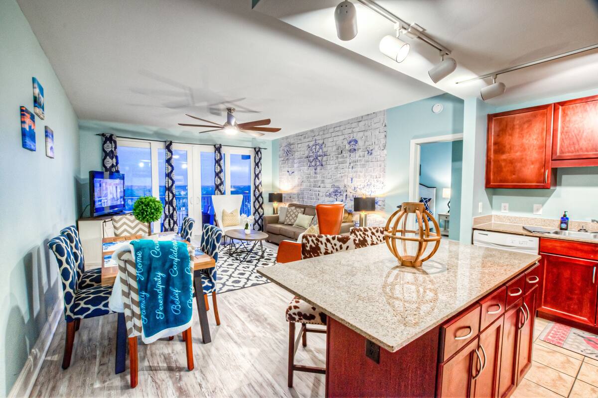 A modern kitchen with a large island, red cabinets, and a dining area with patterned chairs and a blue table runner.
