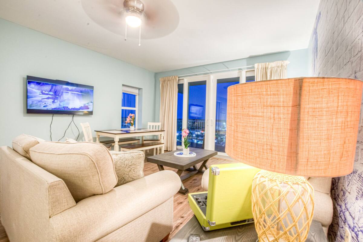 A living room with a sofa, a television, a table with a lamp, and a view of the city at night.