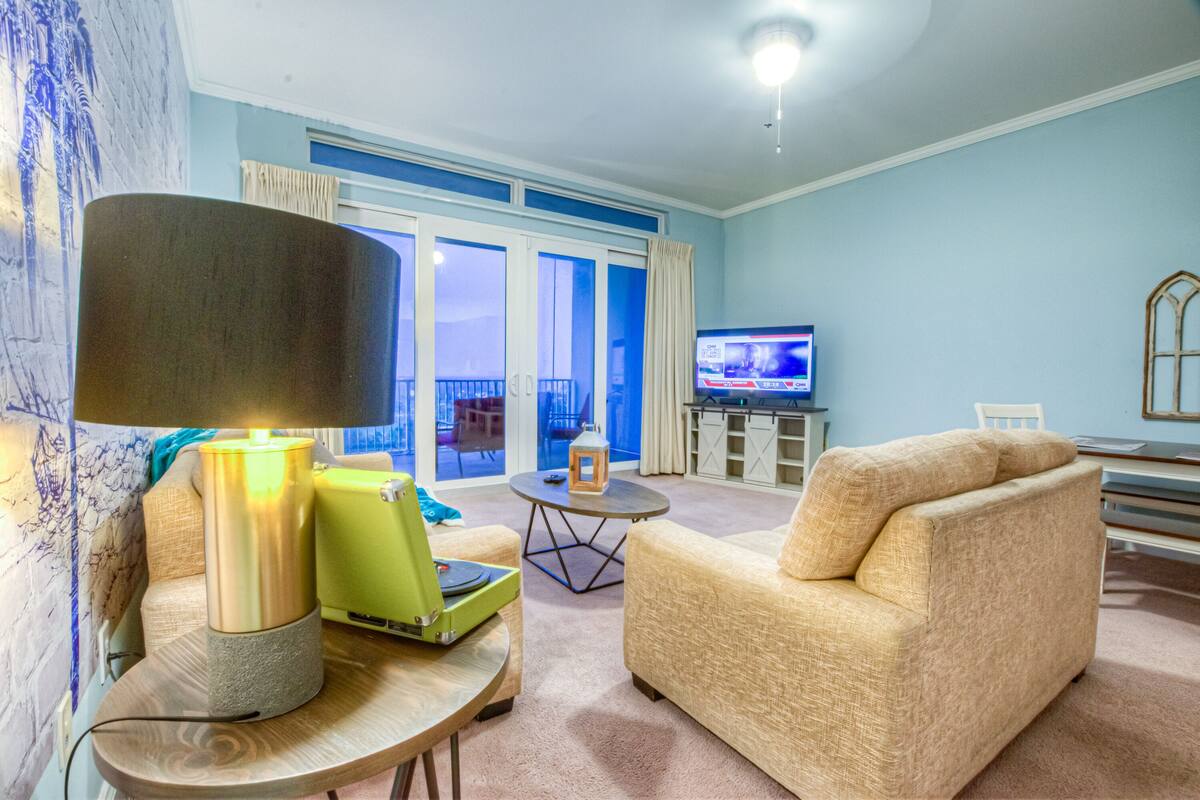 A living room with a sofa, a table with a lamp, a television, and a view of the outside balcony.