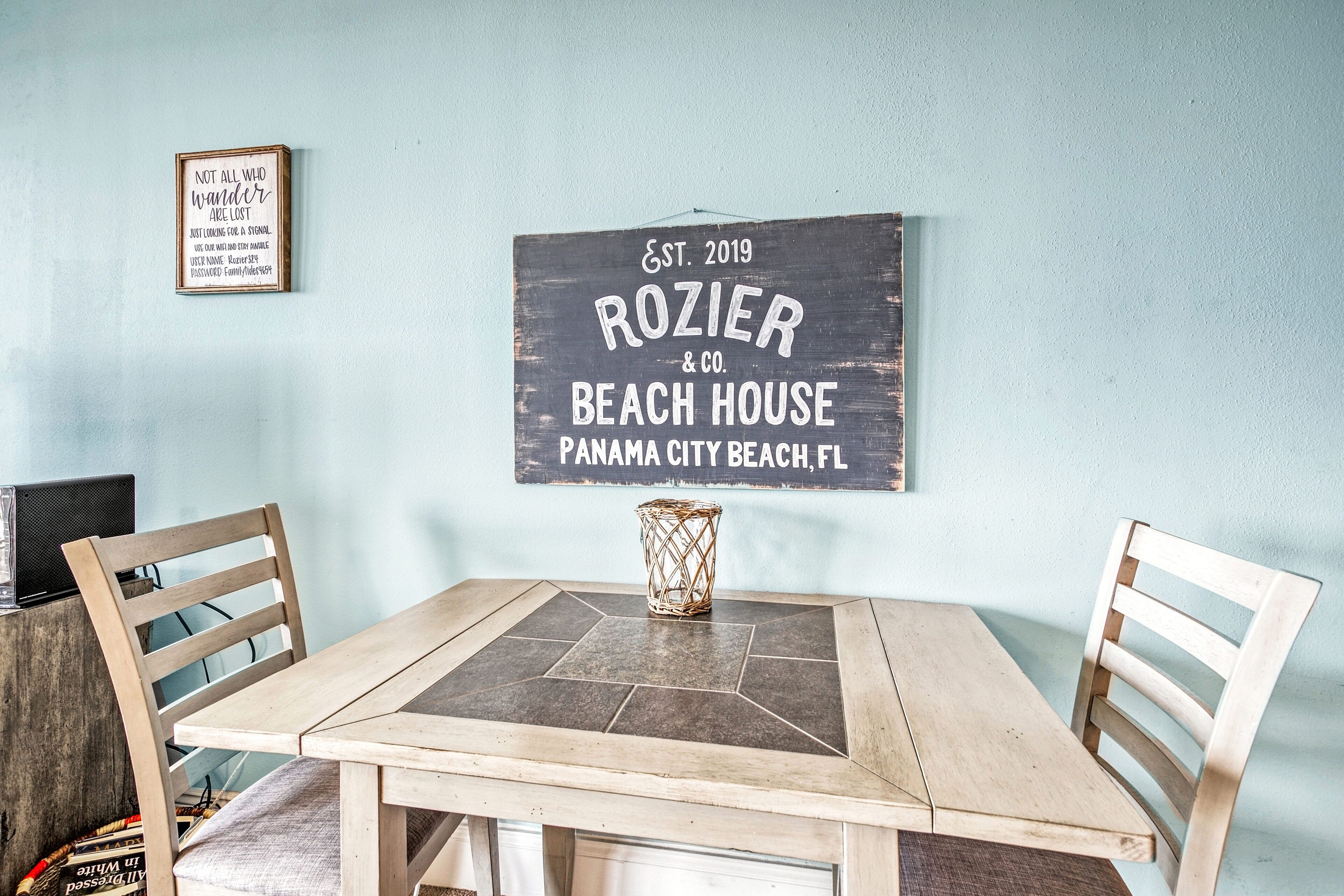 A dining area with a wooden table and chairs, a sign for 'ROZIER & CO. BEACH HOUSE' on the wall, and a decorative vase on the table.