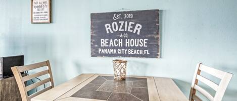 A dining area with a wooden table and chairs, a sign for 'ROZIER & CO. BEACH HOUSE' on the wall, and a decorative vase on the table.