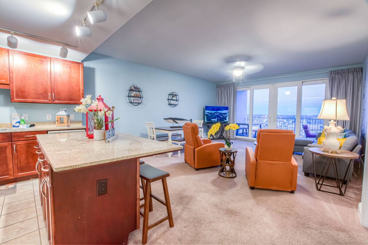 A modern living room with a kitchen area, featuring a large island, seating arrangements, and a television.