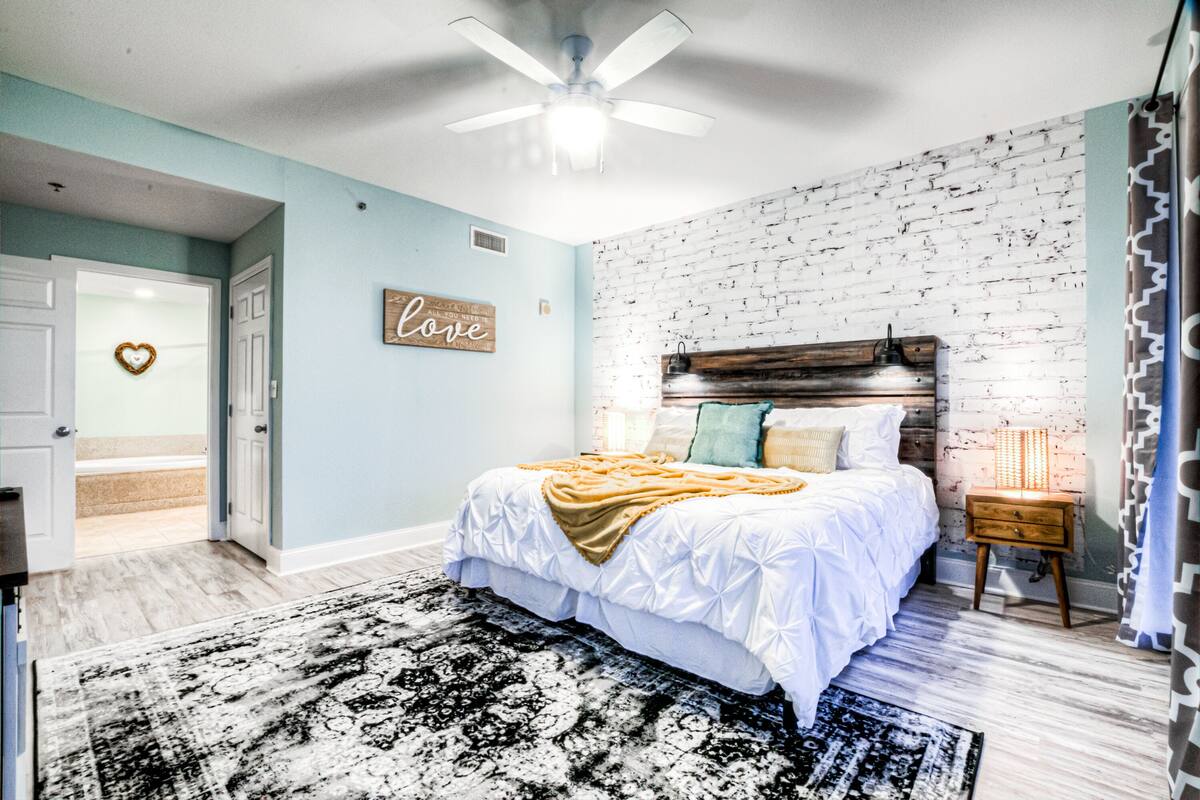 A bedroom with a white bed, a wooden headboard, a black and white rug, a ceiling fan, and a brick wall.
