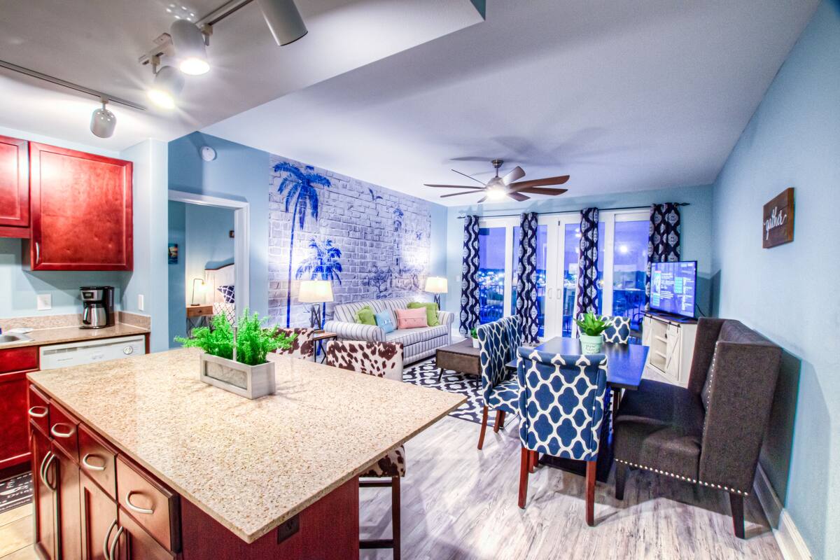A modern kitchen with granite countertops, a dining area with patterned chairs, and a living space with a sofa and a TV.