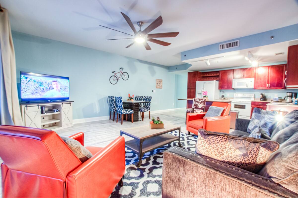 A modern living room with red sofas, a coffee table, and a television. There is a dining area with patterned chairs and a bicycle mounted on the wall. The kitchen has red cabinets and a microwave.