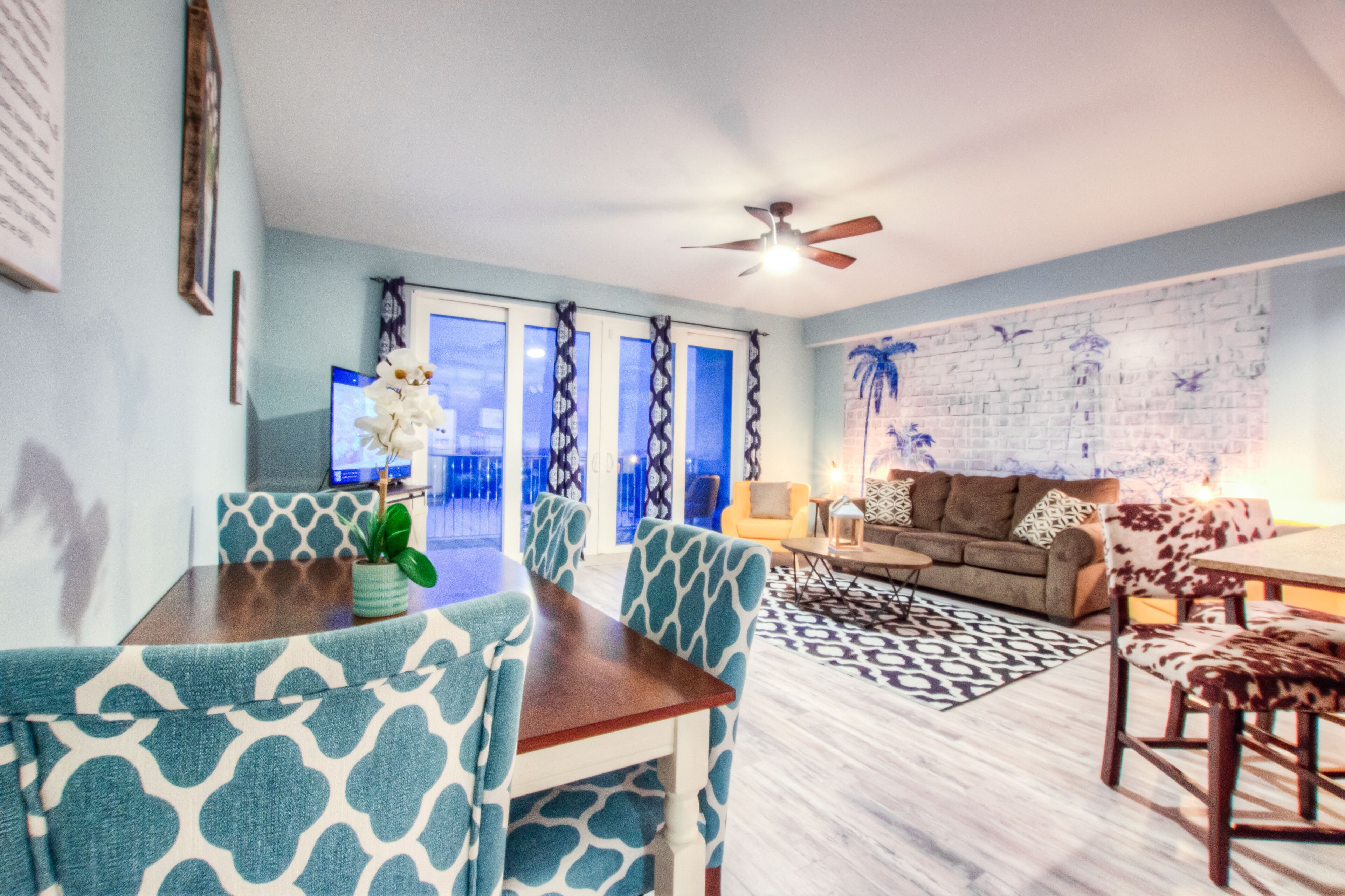 A living room with a dining area, a ceiling fan, a patterned rug, and a wall with a brick design.