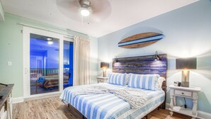 Un dormitorio con una cama grande, un ventilador de techo, una decoración con tabla de surf y un ventanal corredero que da a un balcón.